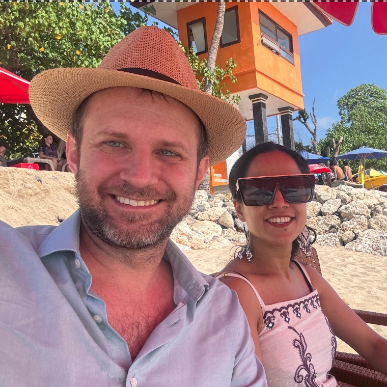 Andrew Long on a beach, in a straw hat and a pale linen shirt, with a companion beside him under a sunny sky.